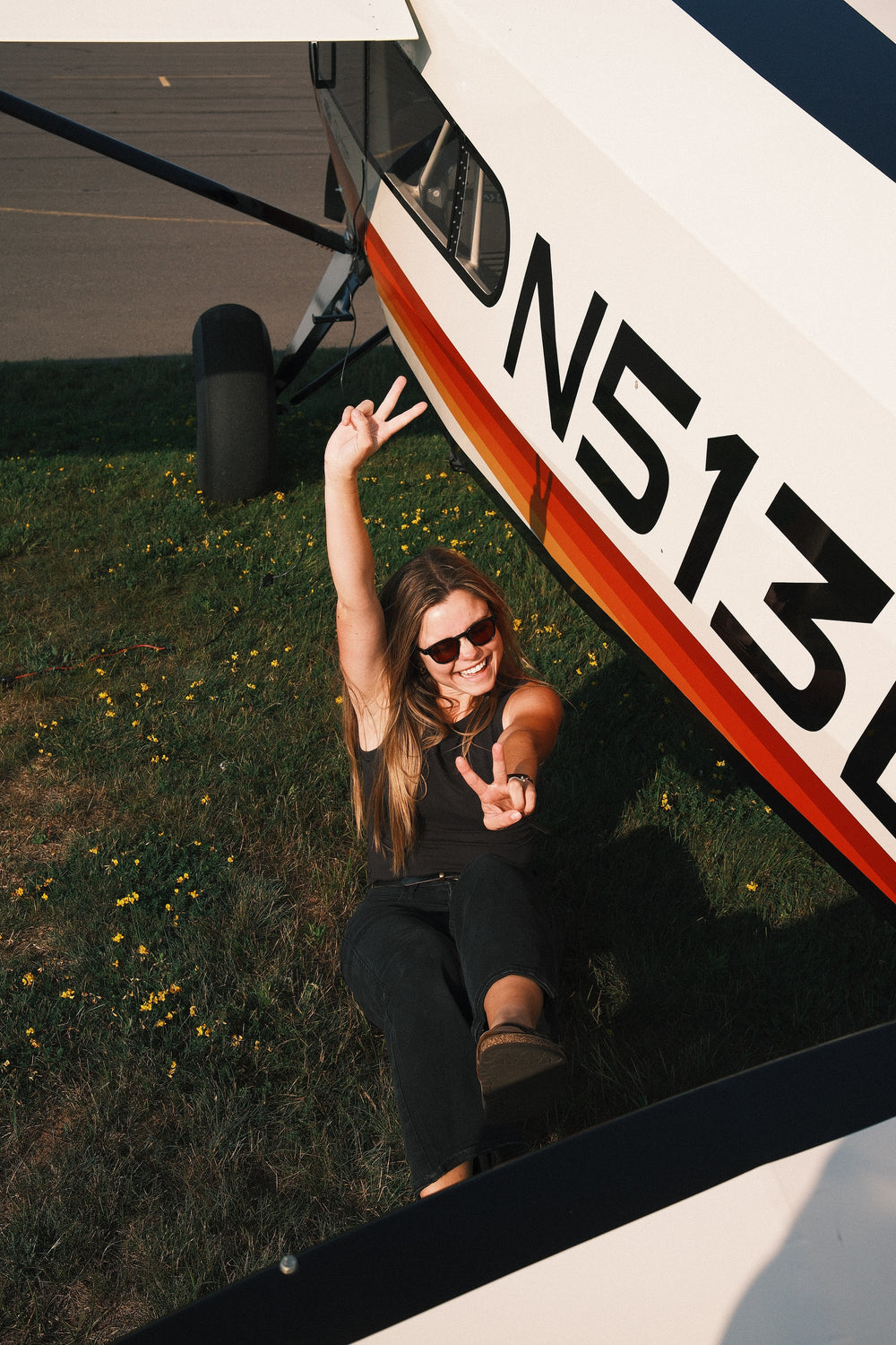 @erinandpaulfly wearing the Kora sunglasses from Otavi Eyewear and a black outfit sits on the grass beside a white airplane, making peace signs toward the camera. The side of the plane shows the registration number DN513. 