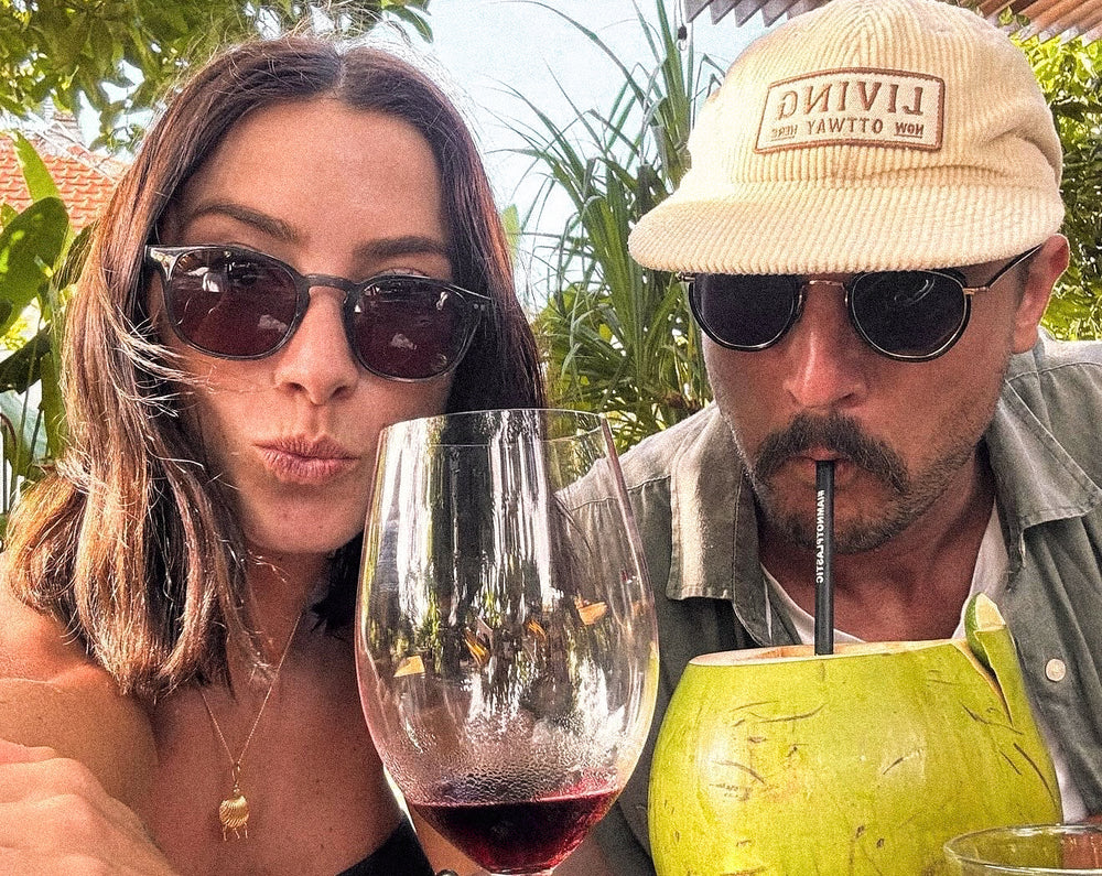 Riley Whitelum and Elayna Karausu posing outdoors with Otavi sunglasses, a glass of red wine, and a coconut drink.